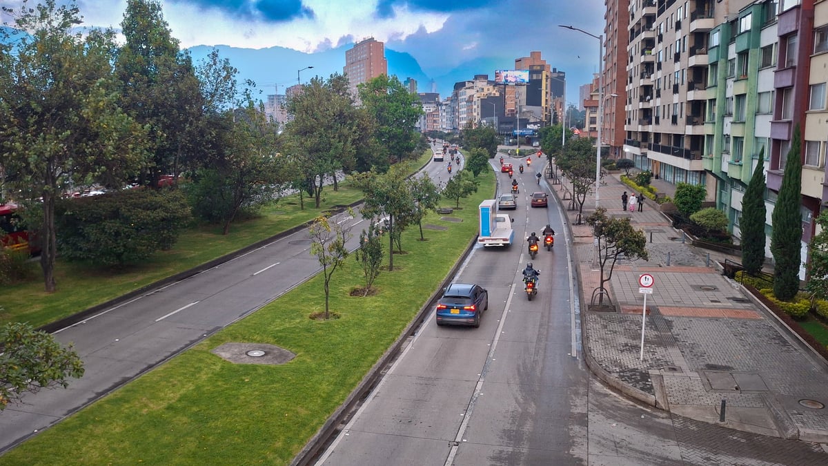 A wide-angle view of a bustling modern avenue in a Latin American city during daylight. The scene features a mix of urban infrastructure and lush green spaces, with cars commuting along the paved road.